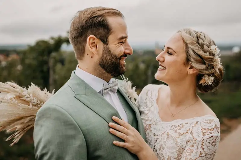 Bräutigam und Braut lächeln sich an, in eleganter Hochzeitskleidung vor natürlicher Kulisse.