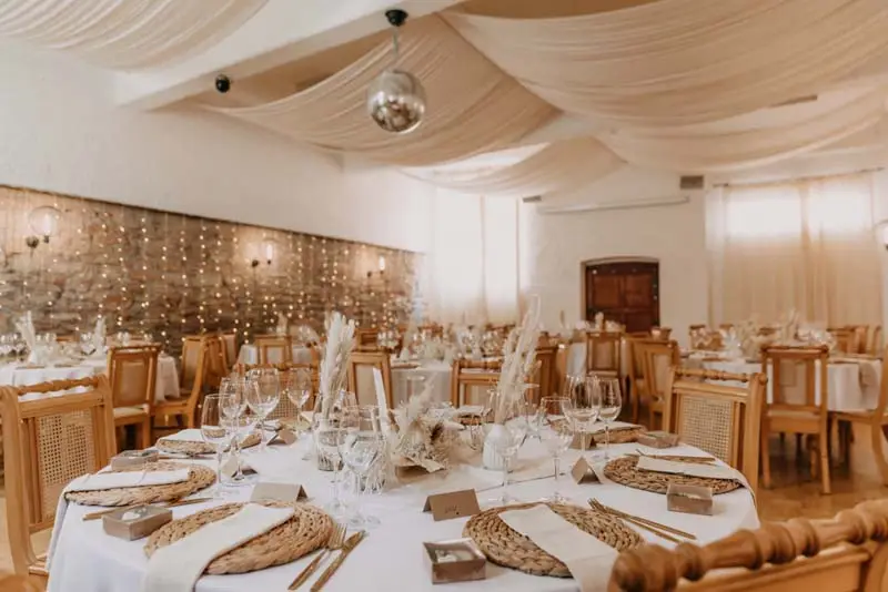 Elegante Hochzeitssaal-Dekoration mit runden Tischen, Glasgeschirr und hängenden Stoffpaneelen in neutralen Tönen.