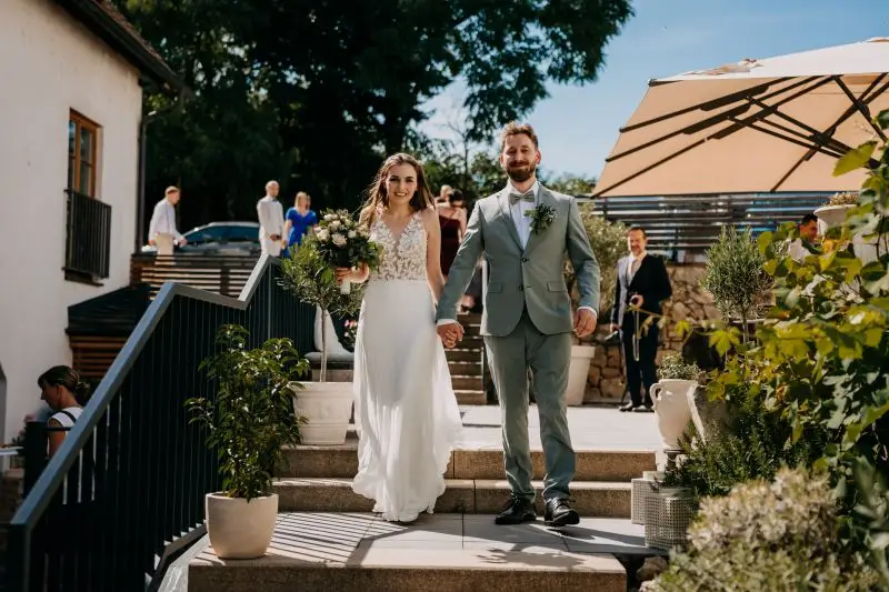 Brautpaar hält Händchen beim Hochzeitsausgang auf sonniger Terrasse mit Gästen und Musik im Hintergrund.