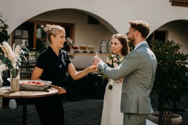 Hochzeitspaar stößt mit Sekt an, während die Hochzeitsplanerin am Tisch mit Snacks lächelt.