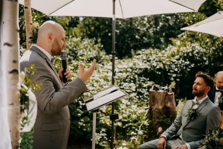 Mann hält Rede auf Outdoor-Hochzeit, während Bräutigam aufmerksam zuhört unter weißen Sonnenschirmen.