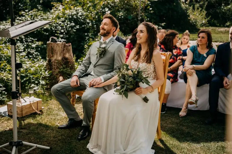 Brautpaar bei einer Gartenhochzeit mit Blumenschmuck und Gästen, die feierlich lächeln und dem Moment folgen.