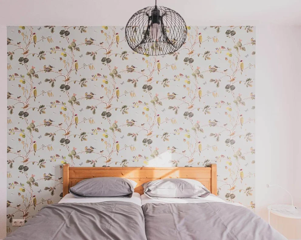 Doppelbett mit grauer Bettwäsche vor floral bedruckter Tapete und moderner Hängelampe im Schlafzimmer.