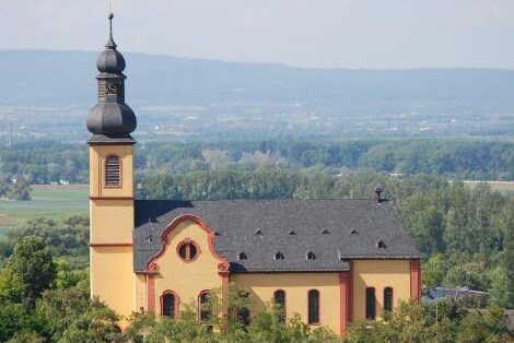 Kleine gelbe Kirche auf einem Hügel mit Wald im Vordergrund und weitem Blick auf das grüne Umland und Berge im Hintergrund.