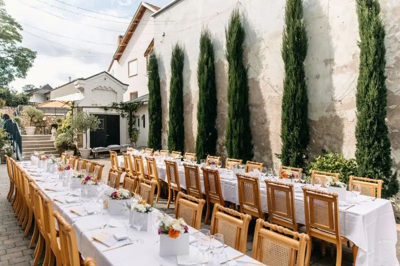 Langer Hochzeitstisch im Freien, umgeben von Zypressen, gedeckt mit weißen Tischdecken und bunten Blumenarrangements.