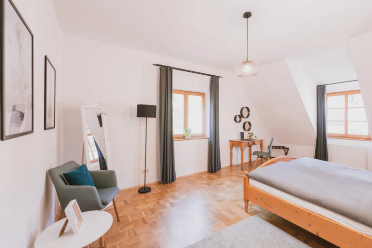 Helles Schlafzimmer mit Holzfußboden, Einzelbett, grauem Sessel, kleinem Tisch, Wanddekoration und Schreibtisch am Fenster.