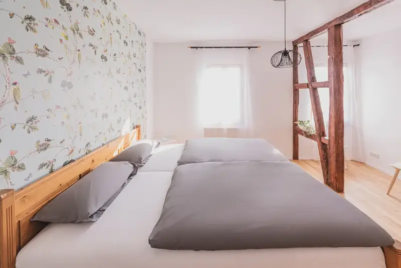 Gemütliches Schlafzimmer mit Doppelbett, floral gemusterter Tapete und Holzbalken in modernem, hellem Design.