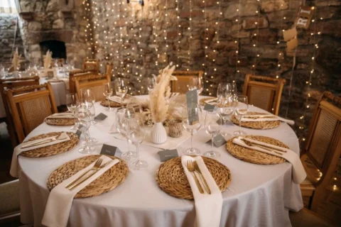 Rund gedeckter Hochzeitstisch mit rustikaler Dekoration, Leinentischdecke, Rattanuntersetzern, Besteck und Gläsern vor gemauerter Wand mit Lichterketten.