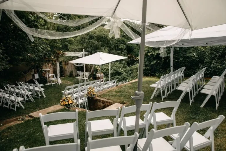 Gartenhochzeit mit weißen Stühlen unter Schirmen, umgeben von grüner Natur und dekoriert mit Blumenarrangements.