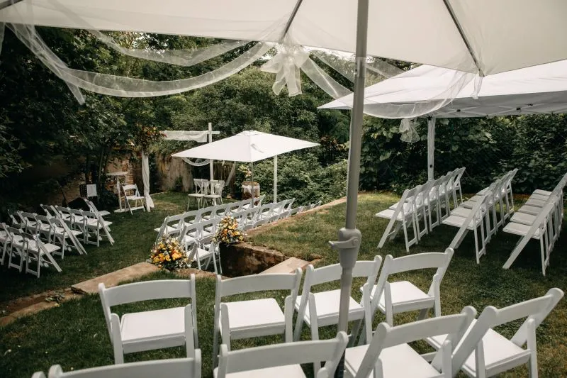 Gartenhochzeit mit weißen Stühlen unter Schirmen, umgeben von grüner Natur und dekoriert mit Blumenarrangements.
