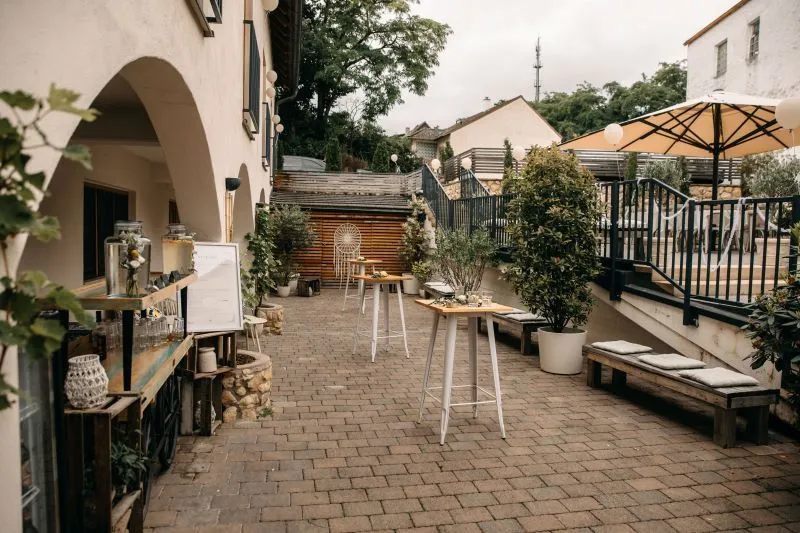 Gemütlicher Außenbereich eines Cafés mit Tischen, Pflanzen und Sitzgelegenheiten auf einer gepflasterten Terrasse.