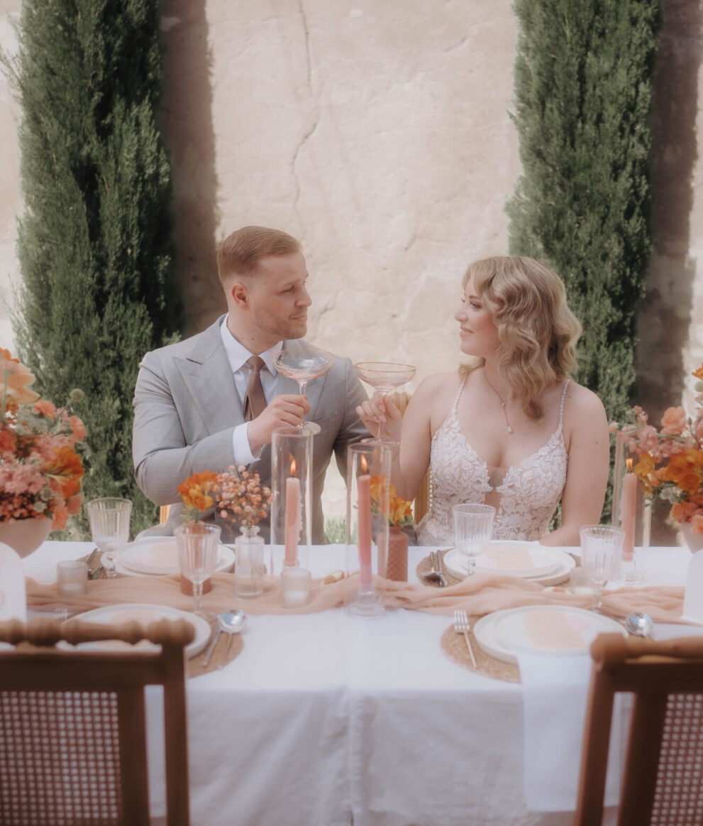 Hochzeitspaar stößt an eleganter Tischdekoration mit Blumen im Freien an, umgeben von mediterraner Kulisse und Zypresse.