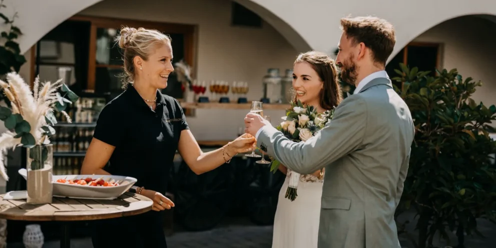 Hochzeitspaar stößt mit Empfangsdame im Freien an, Blumendekoration und Snacks im Hintergrund.