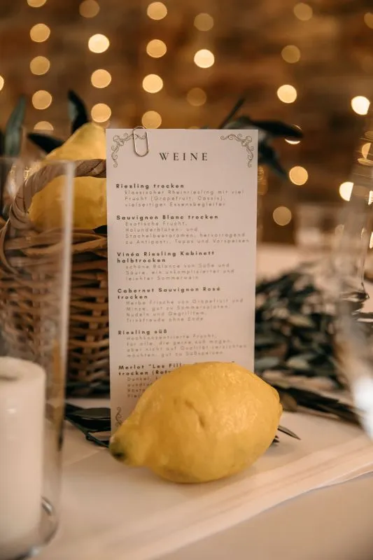 Weinkarte auf Hochzeitstafel mit Zitrone, vor Lichterkette und rustikalem Ambiente.