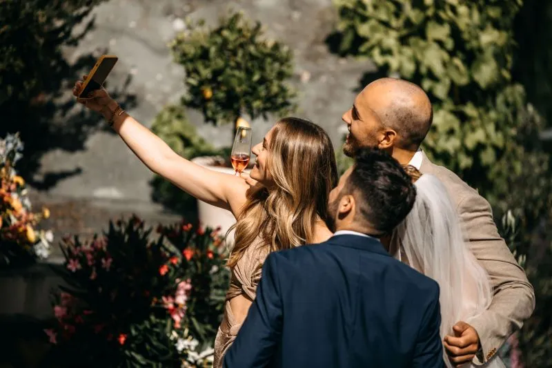 Drei Personen machen ein Selfie auf einer Hochzeit im Freien, umgeben von Blumen und Pflanzen, eine Frau hält ein Glas Sekt.
