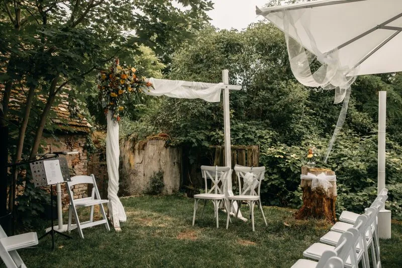 Rustikale Outdoor-Hochzeitsdekoration mit Blumenarrangement und weißen Stühlen im Garten unter einem Baldachin.
