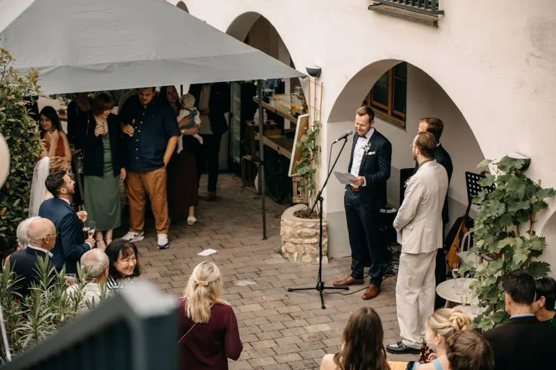 Hochzeitsempfang im Freien mit Redner am Mikrofon und Gästen, die zuhören und sich unterhalten.