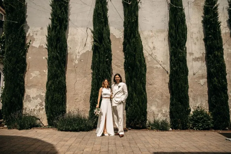 Brautpaar in eleganter Kleidung posiert vor hohen Zypressen; Hochzeit im Freien mit natürlicher Kulisse.