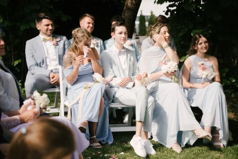 Hochzeit: Junge Gäste in hellblau gekleidet, sitzen draußen auf weißen Stühlen, halten Blumen und wirken festlich.
