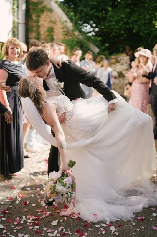 Brautpaar küsst sich romantisch auf Hochzeit mit Blumenstrauß und Gästen im Hintergrund.