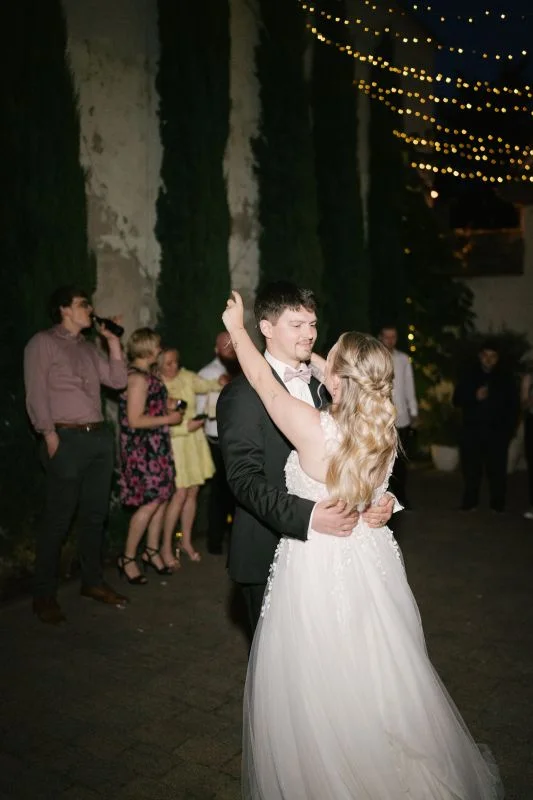 Hochzeitstanz unter Lichterketten im Freien, Braut und Bräutigam lächelnd umarmt von Gästen umgeben