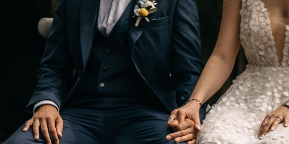 Brautpaar in eleganter Hochzeitskleidung hält Händchen, Detailaufnahme der Hände und Kleidung.