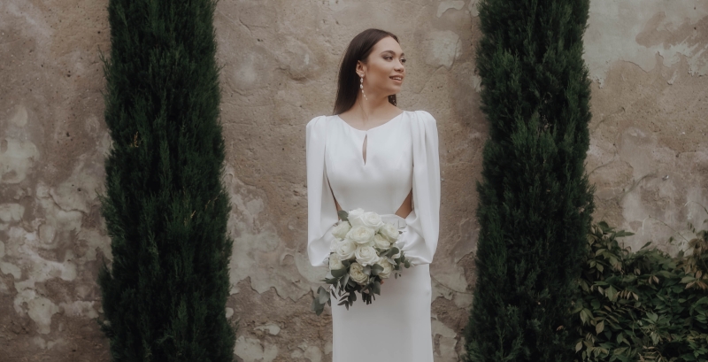 Braut in elegantem Hochzeitskleid mit Blumenstrauß vor rustikaler Steinwand und hochgewachsenen Zypressen.